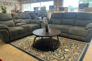 Sala de estar con sofá y loveseat reclinables tapizados en tela gris texturizada, acompañados por una mesa de centro redonda de acabado oscuro sobre una alfombra estampada azul y beige. Exhibición en tienda de muebles.