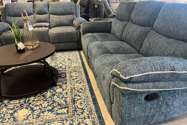 Primer plano de una sala con sofá y loveseat reclinables en tela gris texturizada, mesa de centro redonda de acabado oscuro y alfombra decorativa azul y beige. Se aprecia el mecanismo lateral de reclinación en exhibición de tienda.