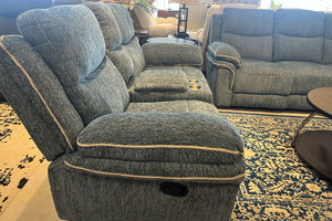 Sofá reclinable de tres plazas y loveseat a juego en tela gris texturizada, con consola central y portavasos integrados. El conjunto se muestra sobre una alfombra azul y beige junto a una mesa de centro redonda en una tienda de muebles.