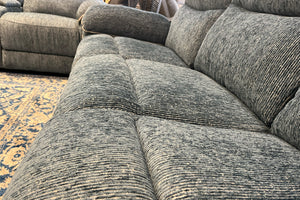 Primer plano de un sofá reclinable de tela gris texturizada con cojines acolchados y asientos amplios. La imagen destaca la suavidad del tapizado y el diseño confortable del mueble en una exhibición de tienda.