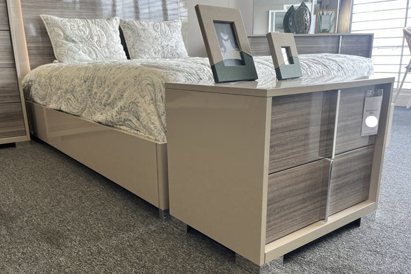 Conjunto de dormitorio moderno que incluye cama en tonos beige con cabecera y estructura en acabado madera, acompañado de una mesa de noche integrada con dos gavetas frontales en acabado madera gris y tiradores verticales metálicos. Sobre la superficie se colocan marcos decorativos, y la cama está vestida con edredón y almohadas en tonos claros con patrón suave. El conjunto se muestra en un ambiente de exhibición con alfombra gris y luz natural.