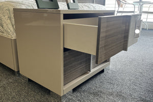 Mesa de noche moderna en acabado beige con frente de gavetas en tono madera gris, mostrando una gaveta parcialmente abierta con interior espacioso en color claro. El mueble presenta líneas rectas, base elevada con soportes transparentes y diseño sin tiradores visibles. Se ubica junto a una cama con ropa de cama en tonos claros y marcos decorativos en la superficie, dentro de un ambiente de exhibición con alfombra gris y luz natural. 