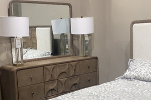 Vista en tienda de gavetero en tono beige con superficies redondeadas y frentes con relieve circular, acompañado de espejo rectangular a juego. Sobre el gavetero hay dos lámparas de mesa con base transparente y pantallas blancas, junto a una cama con ropa de cama en tonos claros al lado.