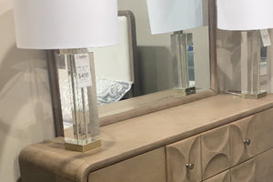 Detalle de gavetero en tono beige con superficie lisa y diseño frontal con relieves circulares, acompañado de espejo rectangular a juego. Sobre el mueble hay dos lámparas de mesa con base transparente y pantalla blanca, en un entorno de tienda.