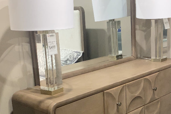 Detalle de gavetero en tono beige con superficie lisa y diseño frontal con relieves circulares, acompañado de espejo rectangular a juego. Sobre el mueble hay dos lámparas de mesa con base transparente y pantalla blanca, en un entorno de tienda.