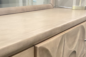 Detalle de superficie superior de gavetero en tono beige con acabado liso y bordes redondeados. Se observa parte de puertas con diseño en relieve curvo y, en la parte posterior, espejo y base de lámpara decorativa en un entorno de tienda.