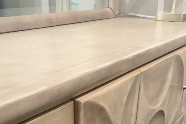 Detalle de superficie superior de gavetero en tono beige con acabado liso y bordes redondeados. Se observa parte de puertas con diseño en relieve curvo y, en la parte posterior, espejo y base de lámpara decorativa en un entorno de tienda.