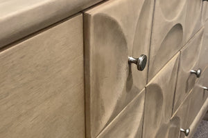 Detalle de gavetero en tono beige con frente de gavetas con relieve curvo y acabado texturizado. Se observa un tirador metálico redondo y la superficie lateral lisa del mueble en un entorno de tienda.