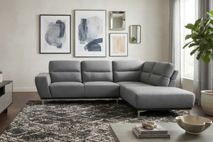 Seccional contemporáneo en tela gris con chaise longue a la derecha y respaldo acolchado. Está ubicado sobre una alfombra de diseño abstracto en una sala moderna con cuadros decorativos, espejos, planta de interior y luz natural proveniente de un amplio ventanal.