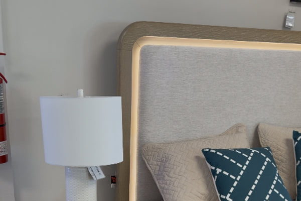 Detalle de cabecera de cama tapizada en tela beige con iluminación LED integrada en el borde, acompañada de cojines decorativos en tonos beige y azul con patrón geométrico y lámpara blanca sobre mesa de noche en un entorno interior.