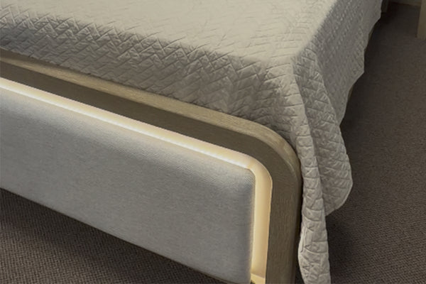 Detalle del pie de cama tapizado en tela beige con bordes redondeados e iluminación LED integrada, acompañado de colcha texturizada en tono claro y alfombra oscura debajo.