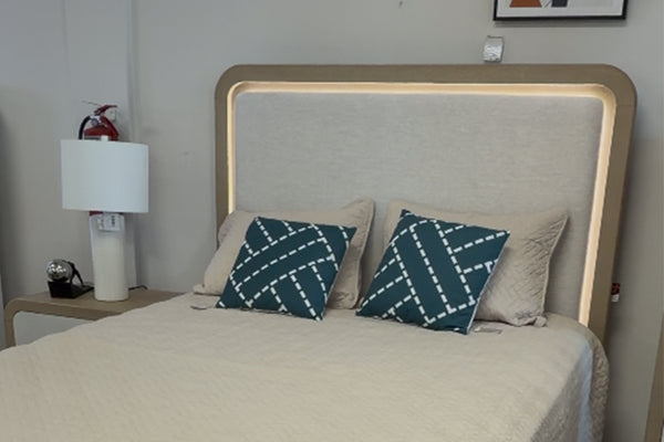 Cabecera de cama moderna tapizada en tela beige con iluminación LED integrada en el marco. La cama está vestida con ropa de cama en tonos neutros y cojines decorativos en azul oscuro con patrón geométrico, acompañada de mesa de noche con lámpara blanca en un entorno interior.