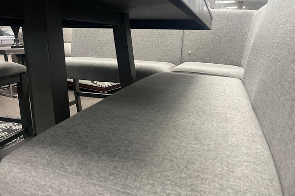 Primer plano de banco tapizado en tela gris claro con respaldo acolchado, integrado a un juego de comedor de altura mostrador. La imagen destaca la textura del tapizado, el amplio asiento y la estructura robusta de la mesa en acabado gris oscuro.