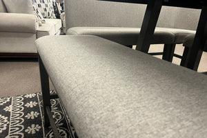 Primer plano de banco tapizado en tela gris claro con asiento acolchado y estructura en acabado gris oscuro. La imagen resalta la textura del tapizado, el diseño limpio y la integración con un juego de comedor contemporáneo de altura mostrador.