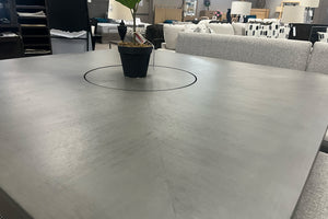 Primer plano de mesa de comedor de altura mostrador en acabado gris con superficie amplia y diseño contemporáneo. En el centro se aprecia una bandeja giratoria integrada con un arreglo decorativo, destacando la textura y funcionalidad de la cubierta.