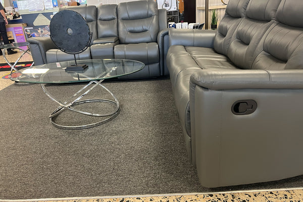 Sala moderna con sofá y loveseat reclinables en color gris oscuro, tapizados en material tipo cuero con cojines acolchados y respaldos altos. El conjunto está acompañado por una mesa de centro de vidrio con base metálica plateada y decoración contemporánea en un showroom de muebles.