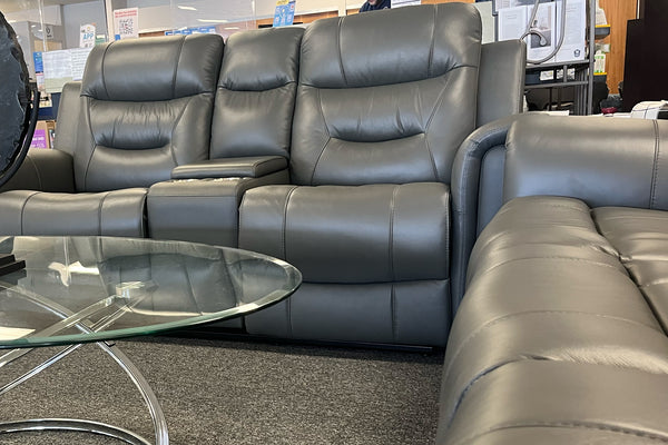 Loveseat reclinable en color gris oscuro con tapizado tipo cuero, respaldos altos acolchados y consola central con apoyabrazos integrados. La imagen muestra parte de un sofá a juego y una mesa de centro de vidrio con base metálica en un showroom de muebles modernos.