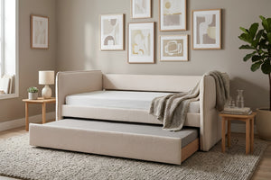 Cama tipo daybed tapizada en tela beige texturizada con cama nido inferior extraíble. Presentada en un dormitorio de estilo contemporáneo con mesas auxiliares de madera, arte mural abstracto y decoración en tonos neutros.