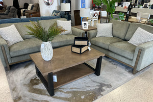 Juego de sala contemporáneo con sofá y loveseat tapizados en tela verde oliva, cojines decorativos texturizados en color crema y detalles de madera en los brazos, acompañado de una mesa de centro de madera con base negra y decoración floral, exhibido en una tienda de muebles moderna.