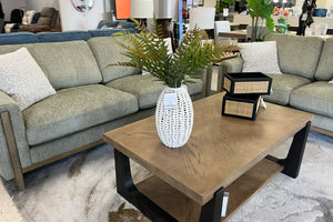 Juego de sala contemporáneo con sofá y loveseat tapizados en tela verde oliva, cojines decorativos color crema y mesa de centro de madera con base negra, decorada con un florero blanco con hojas verdes y cajas decorativas, exhibido en una tienda de muebles moderna.