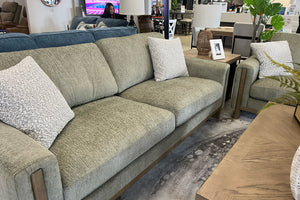 Sofá contemporáneo tapizado en tela verde oliva con cojines decorativos texturizados en color crema, brazos rectos con detalles de madera y asientos amplios acolchados, exhibido junto a un sillón a juego en una tienda de muebles moderna.