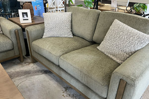 Loveseat contemporáneo tapizado en tela verde oliva con cojines decorativos texturizados en color crema, brazos rectos con detalles de madera y asiento acolchado, exhibido en una tienda de muebles sobre una alfombra de diseño abstracto.