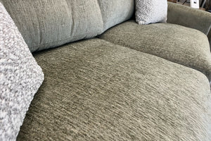 Primer plano de un loveseat tapizado en tela verde oliva, mostrando los asientos acolchados y la textura tejida de la tela, con cojines decorativos suaves en color crema en una sala de exhibición de muebles.
