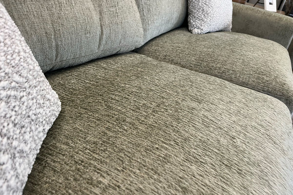 Primer plano de un loveseat tapizado en tela verde oliva, mostrando los asientos acolchados y la textura tejida de la tela, con cojines decorativos suaves en color crema en una sala de exhibición de muebles.