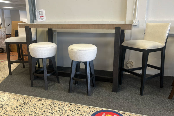Juego de comedor alto con mesa rectangular de madera clara y patas negras, acompañado de dos bancos redondos tapizados en tela blanca y dos sillas altas acolchadas en color beige, exhibido en una tienda de muebles sobre alfombra gris.