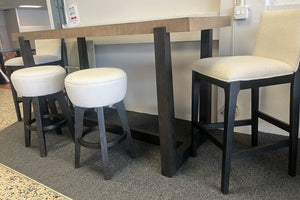 Mesa alta rectangular de madera clara con estructura negra, acompañada de dos bancos redondos tapizados en tela blanca y sillas altas acolchadas en color crema, exhibida en una tienda de muebles sobre alfombra gris.