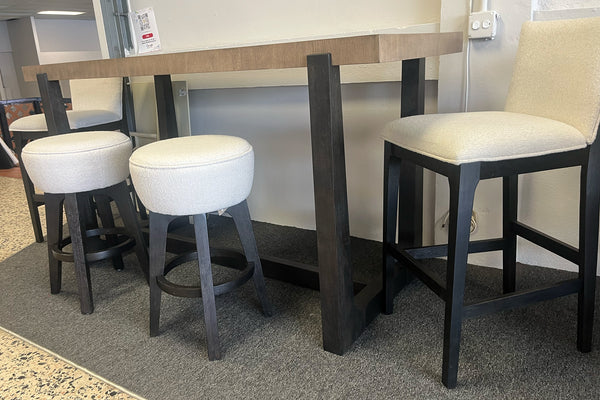 Mesa alta rectangular de madera clara con estructura negra, acompañada de dos bancos redondos tapizados en tela blanca y sillas altas acolchadas en color crema, exhibida en una tienda de muebles sobre alfombra gris.