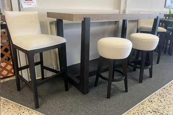 Juego de comedor alto con mesa rectangular de madera clara y patas negras, acompañado de dos bancos redondos tapizados en tela blanca y sillas altas acolchadas en color crema, exhibido en una tienda de muebles sobre alfombra gris oscura.