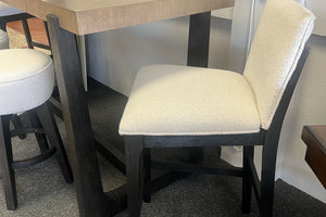 Silla alta contemporánea tapizada en tela color crema con estructura de madera negra, colocada junto a una mesa alta de madera clara y un banco redondo a juego, exhibida en una tienda de muebles moderna.