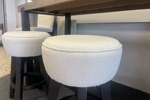 Dos bancos altos redondos tapizados en tela blanca texturizada con patas de madera oscura, colocados debajo de una mesa alta de madera y metal en una exhibición de tienda de muebles moderna.