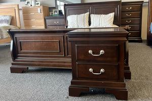 Juego de dormitorio de estilo clásico en madera color marrón oscuro, compuesto por cama con cabecera y piecera con paneles decorativos, y mesa de noche con dos gavetas y tiradores metálicos curvos. En el fondo se observan un chest a juego y almohadas sobre la cama, todo expuesto en una sala de exhibición con alfombra gris.
