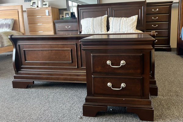 Juego de dormitorio de estilo clásico en madera color marrón oscuro, compuesto por cama con cabecera y piecera con paneles decorativos, y mesa de noche con dos gavetas y tiradores metálicos curvos. En el fondo se observan un chest a juego y almohadas sobre la cama, todo expuesto en una sala de exhibición con alfombra gris.