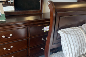 Gavetero hecha de madera marrón oscuro con seis gavetas y con tiradores metálicos detrás de una cama al mismo diseńo con acolchado blanco, todo en un fondo de tienda. 