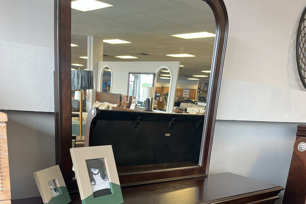 Espejo en marco marrón oscuro con esquinas redondeadas y objetos decorativos encima de gavetero, todo en un fondo de tienda. 