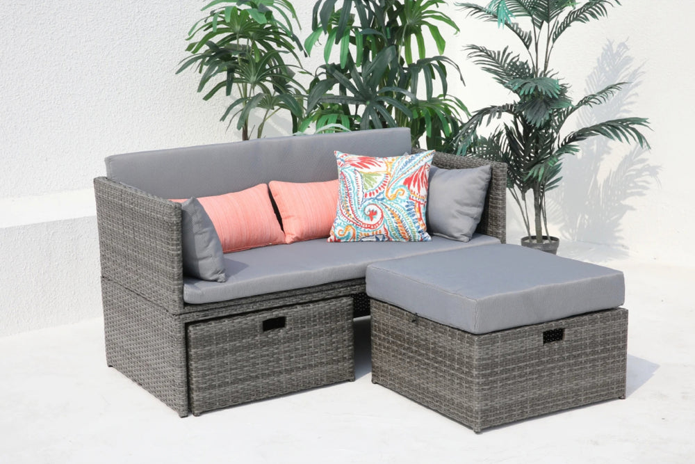 Sofá de ratán para exterior con cojines grises, reposapiés y almohadones decorativos con plantas.