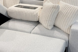 Primer plano de un sofá modular tapizado en tela blanca texturizada con chaise longue, cojines decorativos acanalados y consola central acolchada con portavasos integrados en acabado negro.