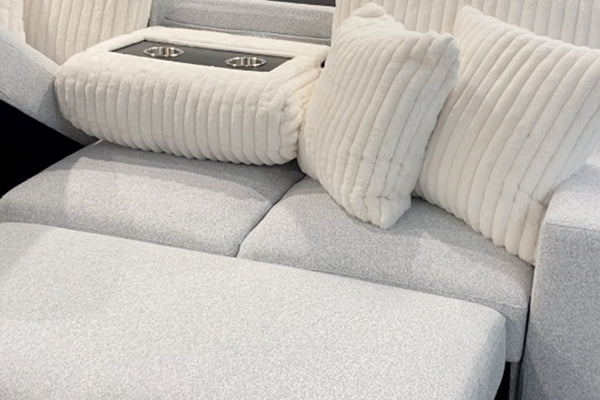 Primer plano de un sofá modular tapizado en tela blanca texturizada con chaise longue, cojines decorativos acanalados y consola central acolchada con portavasos integrados en acabado negro.