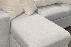 Primer plano de un sofá modular contemporáneo con chaise longue tapizada en tela blanca texturizada, acompañado de cojines decorativos acanalados en color crema y asientos acolchados de gran tamaño.