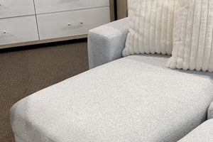 Primer plano de una chaise longue modular tapizada en tela blanca texturizada, con cojines decorativos acanalados en color crema y diseño acolchado contemporáneo, exhibida en una tienda de muebles moderna.