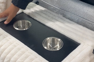 Consola central acolchada de sofá modular tapizado en tela blanca tipo felpa, con dos portavasos metálicos integrados y superficie negra para colocar bebidas o accesorios.