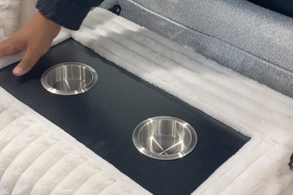 Consola central acolchada de sofá modular tapizado en tela blanca tipo felpa, con dos portavasos metálicos integrados y superficie negra para colocar bebidas o accesorios.