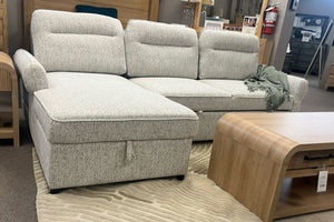 Sofá seccional con chaise en tela color marfil jaspeado, respaldos acolchados y compartimento de almacenamiento integrado en la chaise. Exhibido en una sala de muebles junto a una mesa de centro de madera con diseño curvo.