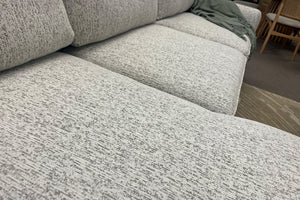 Primer plano de la tela jaspeada en tonos marfil y gris de un sofá seccional con chaise. La imagen destaca la textura del tapizado, los cojines acolchados y la amplitud de los asientos para mayor comodidad.
