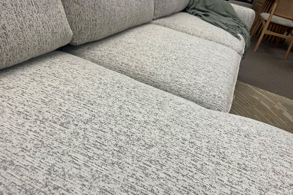 Primer plano de la tela jaspeada en tonos marfil y gris de un sofá seccional con chaise. La imagen destaca la textura del tapizado, los cojines acolchados y la amplitud de los asientos para mayor comodidad.