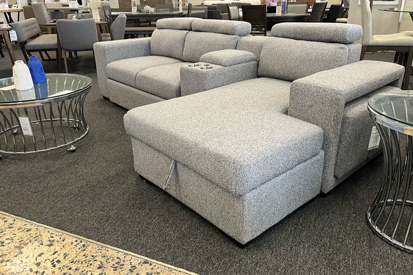 Sofá seccional gris claro con chaise longue, reposacabezas acolchados ajustables y consola central con portavasos. La imagen muestra el mueble exhibido en una tienda de muebles, rodeado de mesas auxiliares con cubierta de vidrio.