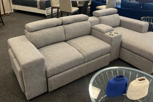 Loveseat moderno en color gris claro con tapizado texturizado, reposacabezas ajustables y consola central con dos portavasos. Exhibido en una tienda de muebles junto a una chaise longue a juego y una mesa auxiliar con cubierta de vidrio.